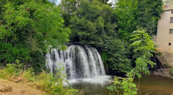 Belle cascade auvergne, entourée de verdure luxuriante. L'eau tombe en rideau sur des rochers, rejoignant une rivière paisible. Un bâtiment ancien se dresse à droite.