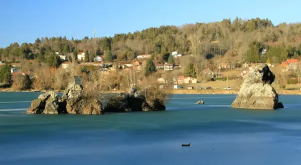 Le lac de Cassière gelé, avec ses îlots rocheux et ses maisons sur la colline boisée, sous un ciel bleu clair.