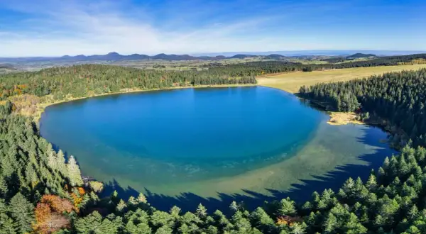 Vue aérienne du lac de Servières, entouré de forêts verdoyantes et de montagnes bleutées sous un ciel clair.