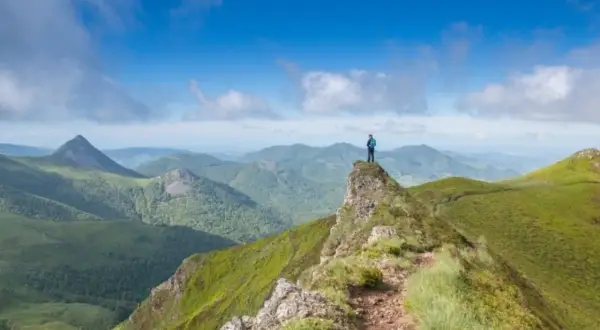 Un randonneur au sommet d'une crête verdoyante, contemplant un panorama de montagnes. Quel est le plus beau coin du Cantal ? Ce paysage spectaculaire en est un candidat.