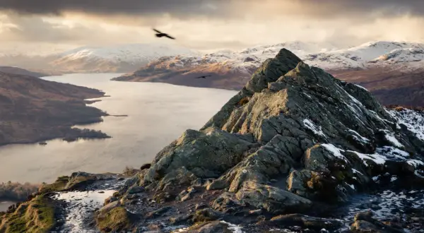 Vue spectaculaire d'une montagne écossaise enneigée, avec un lac et des oiseaux en vol.