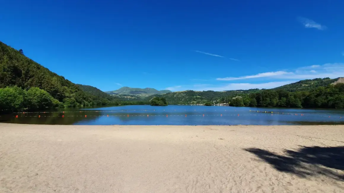 Plage de sable fin au bord d'un lac paisible, avec des montagnes verdoyantes en arrière-plan, sous un ciel bleu éclatant. Le Puy de Sancy veille sur ce paysage idyllique.