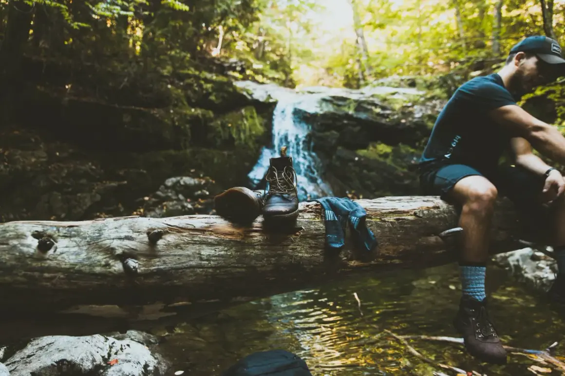 Un randonneur se repose près d'une cascade, ses bottes et chaussettes posées sur une bûche. Une pause pour réfléchir à comment éviter de transpirer des pieds.
