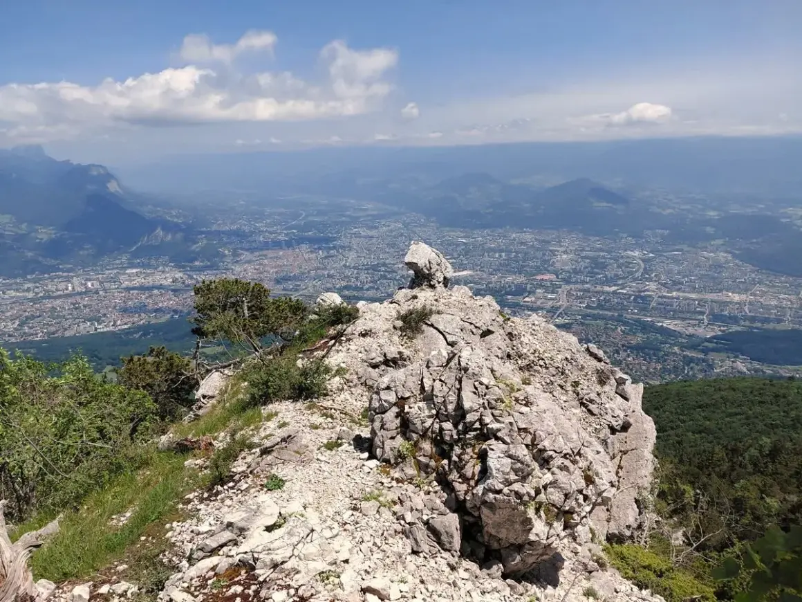 Vue panoramique depuis le sommet rocheux du Moucherotte, dominant une ville étendue sous un ciel bleu parsemé de nuages.