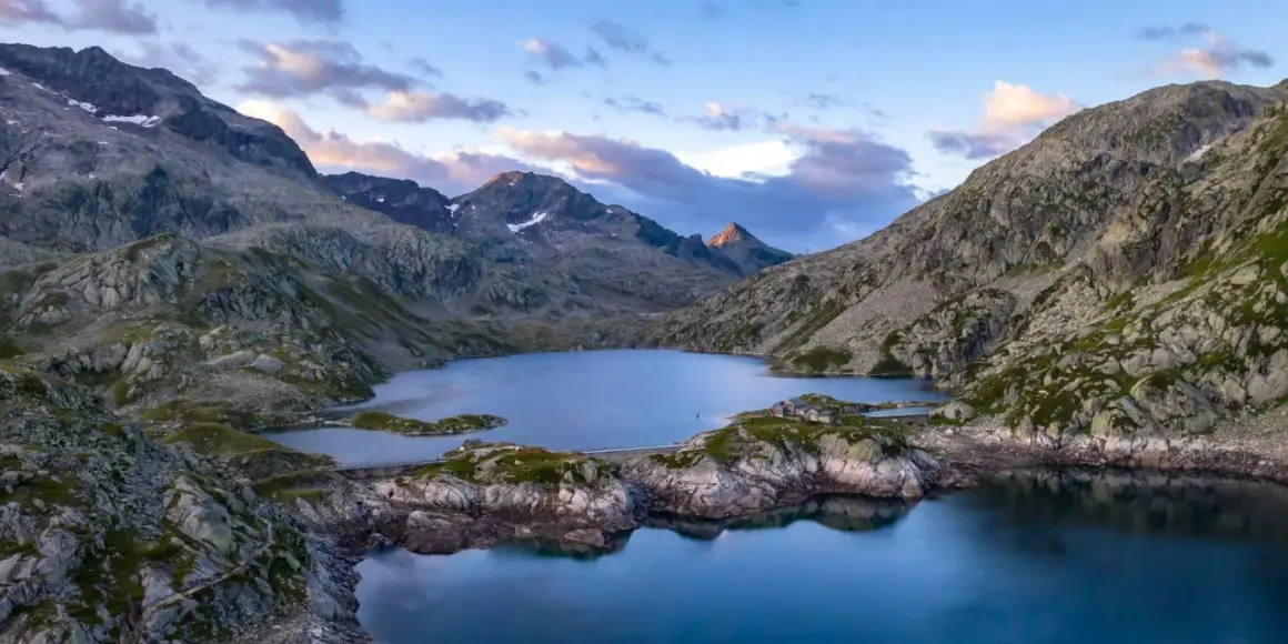 Vue aérienne du lac des 7 Laux, entouré de montagnes rocheuses et verdoyantes, sous un ciel aux teintes rosées.