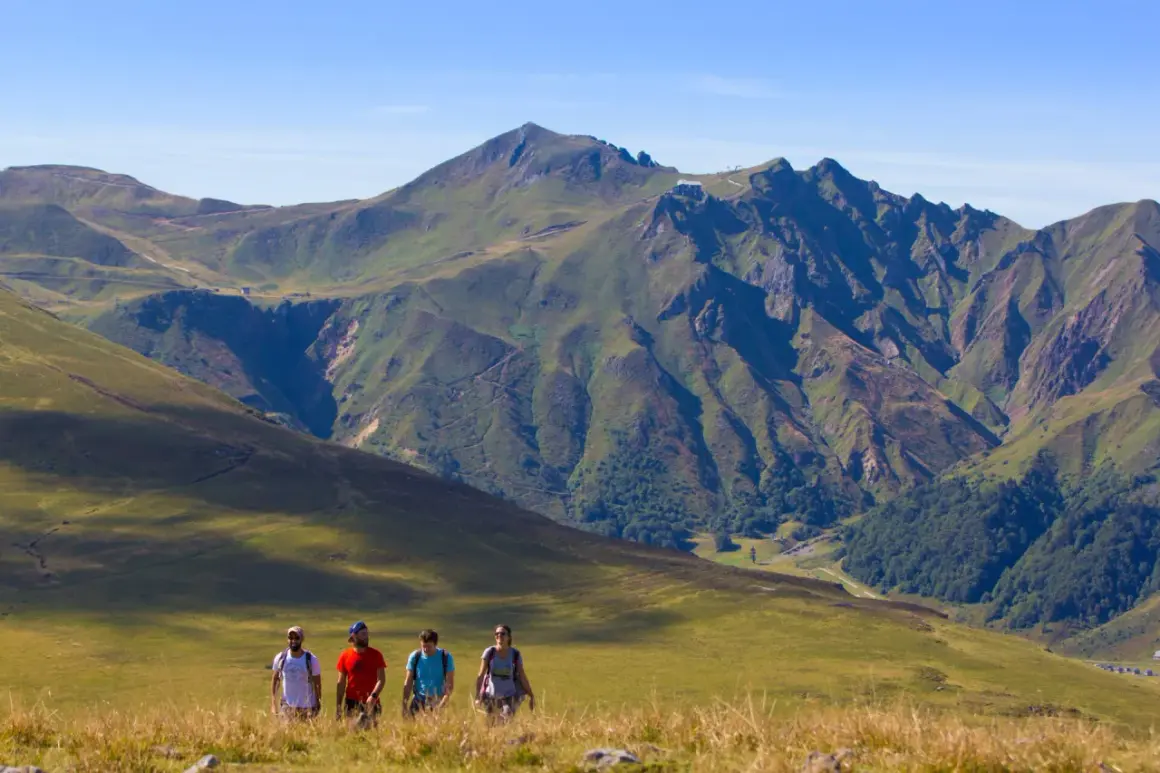 Quatre randonneurs marchent dans un paysage de montagne verdoyant, avec le plus haut sommet du Massif Central en toile de fond.