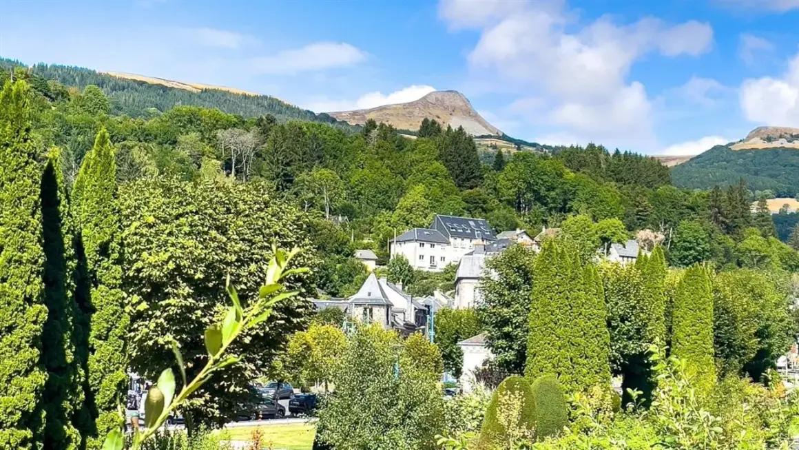 Vue d'un village niché dans une vallée verdoyante, avec des maisons aux toits gris et un grand bâtiment blanc. Au loin, une montagne imposante se dresse sous un ciel bleu. Un banne ordanche, peut-être une tente ou un auvent, est visible dans le paysage.