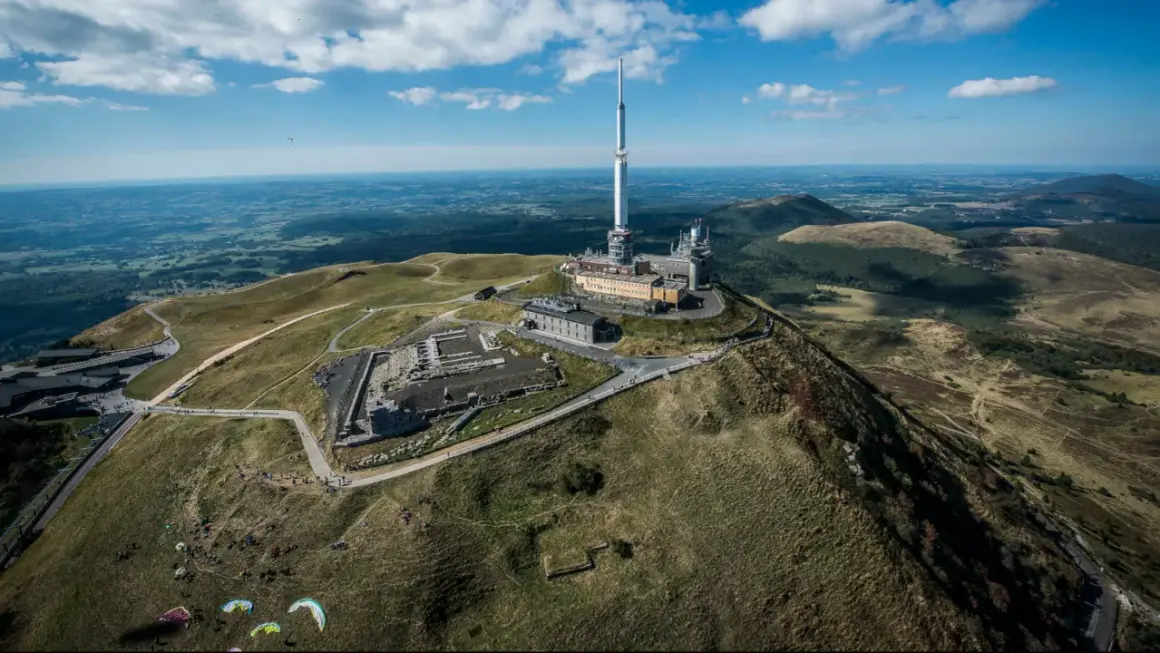 Vue aérienne du Puy de Dôme dans le Massif Central, avec sa tour de télécommunication et des parapentistes profitant du paysage.