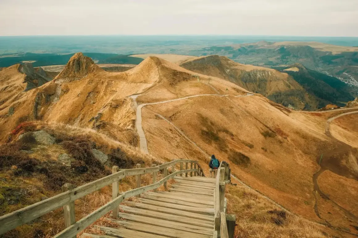 Une personne entame une rando au Puy de Sancy, descendant un escalier en bois sur un flanc de montagne aux herbes dorées.