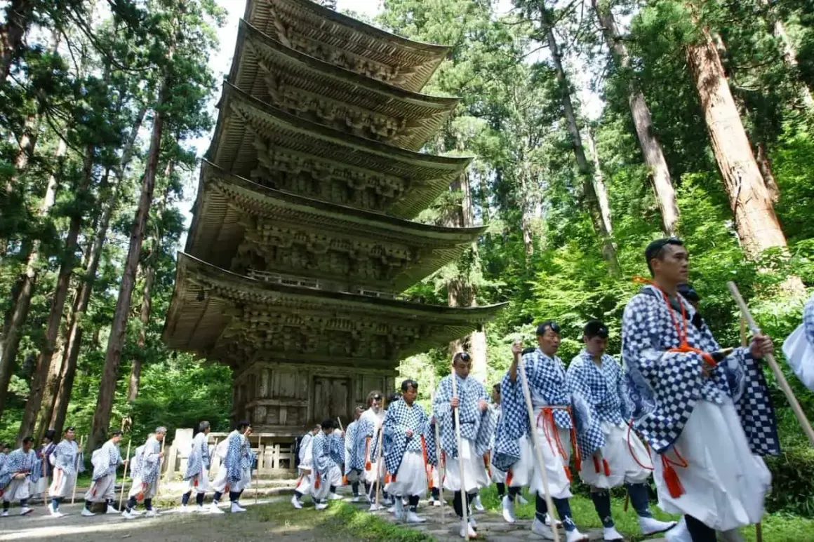 Marche d'hommes en tenue traditionnelle, portant des bâtons, devant une pagode à cinq étages, dans le cadre d'une cérémonie Dewa Sanzan.