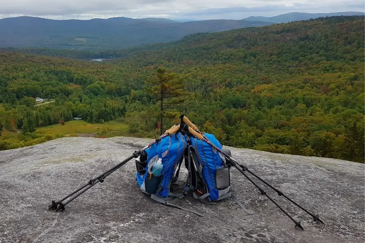 Deux sacs de rando bleus sont posés sur un rocher, avec des bâtons de marche plantés à côté. Le paysage vallonné et boisé s'étend à l'horizon.