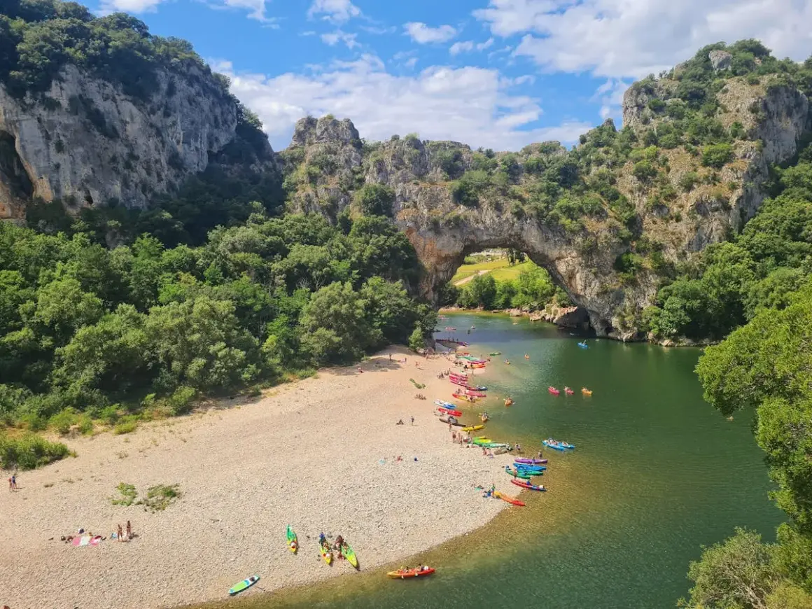Les gorges du Chassezac, un pont naturel rocheux surplombe une rivière verdoyante où des kayakistes profitent du soleil sur une plage de galets.