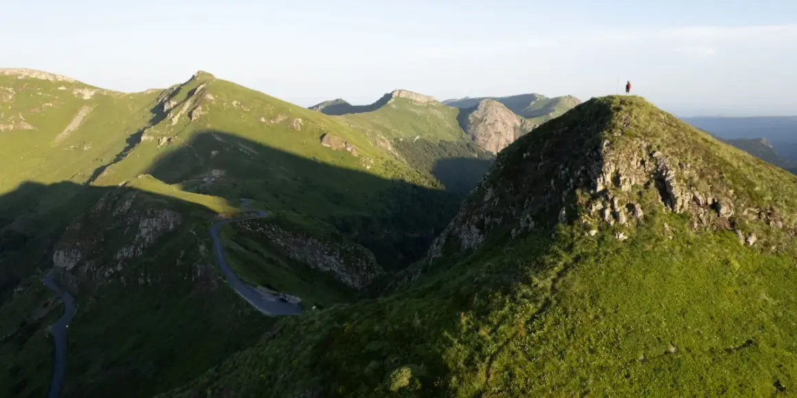 Un randonneur solitaire au sommet d'une colline verdoyante, dominant un paysage montagneux spectaculaire. Une route sinueuse serpente dans la vallée. Randonnée Cantal.