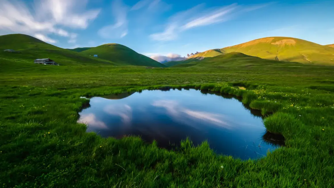 Un étang reflète le ciel et les collines verdoyantes, évoquant la petite Mongolie auvergnate. Un paysage paisible avec des nuages filant.