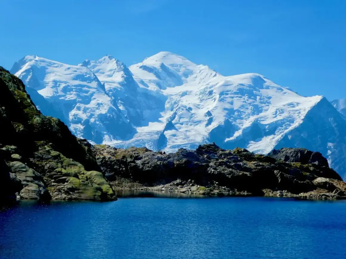 Lac d'altitude aux eaux bleues, bordé de roches sombres, avec en arrière-plan le massif enneigé du Mont-Blanc, paysage typique du trek des Aiguilles Rouges.