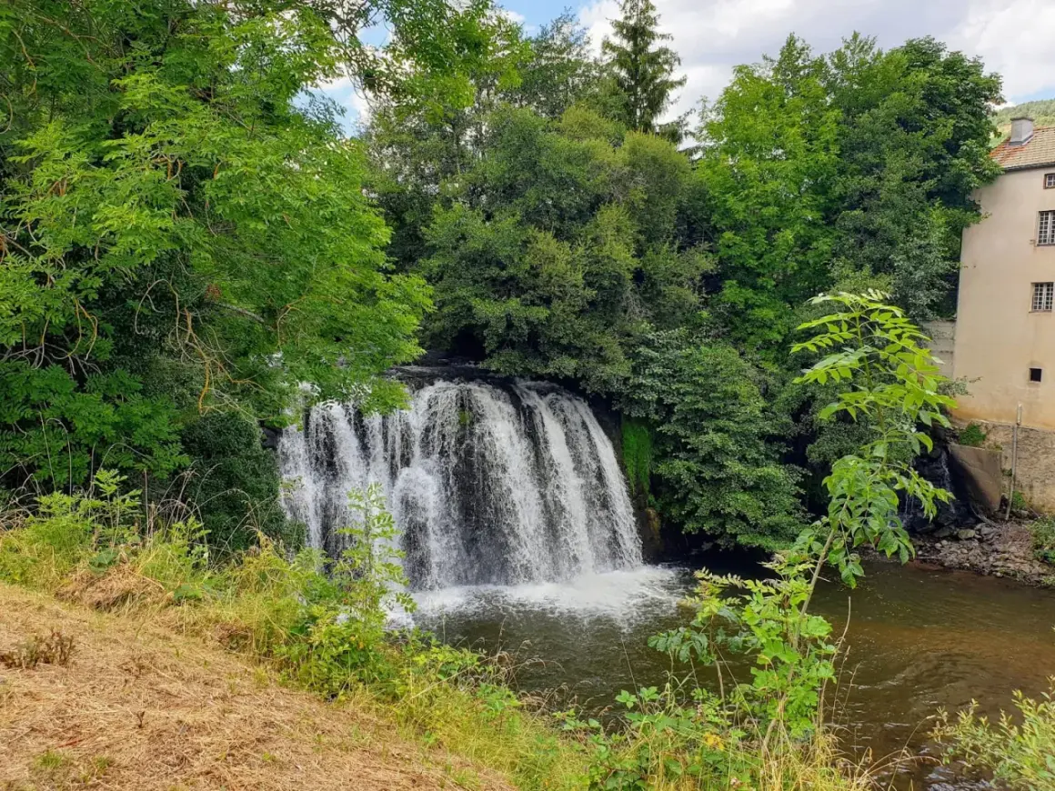 Belle cascade auvergne, entourée de verdure luxuriante. L'eau tombe en rideau sur des rochers, rejoignant une rivière paisible. Un bâtiment ancien se dresse à droite.