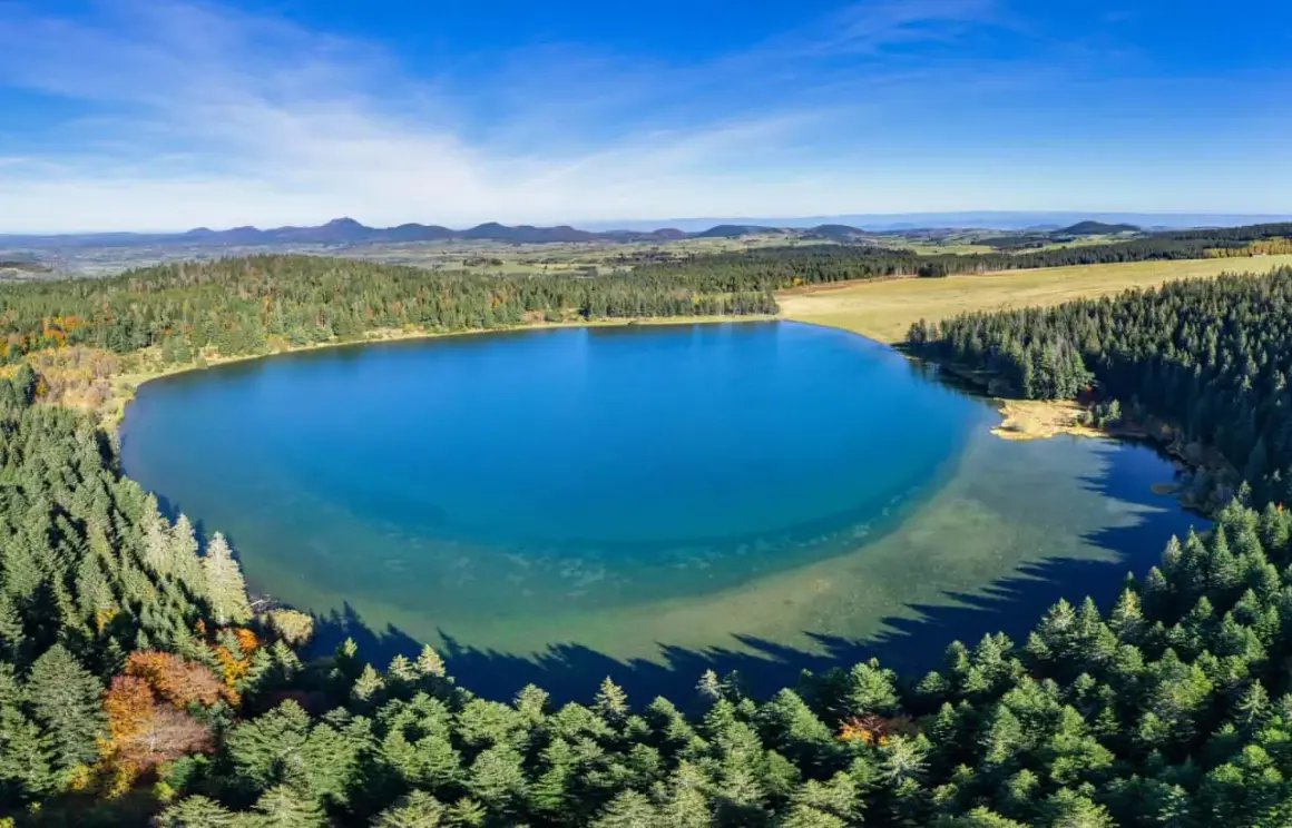 Vue aérienne du lac de Servières, entouré de forêts verdoyantes et de montagnes bleutées sous un ciel clair.