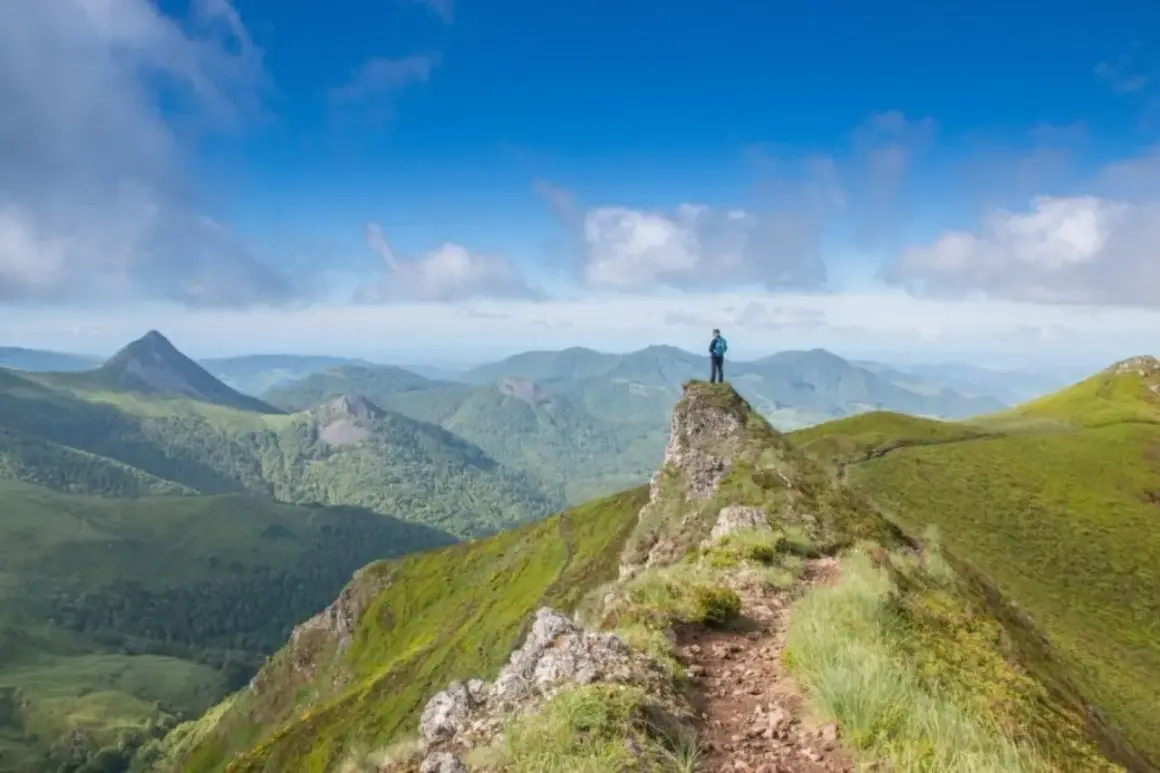 Un randonneur au sommet d'une crête verdoyante, contemplant un panorama de montagnes. Quel est le plus beau coin du Cantal ? Ce paysage spectaculaire en est un candidat.
