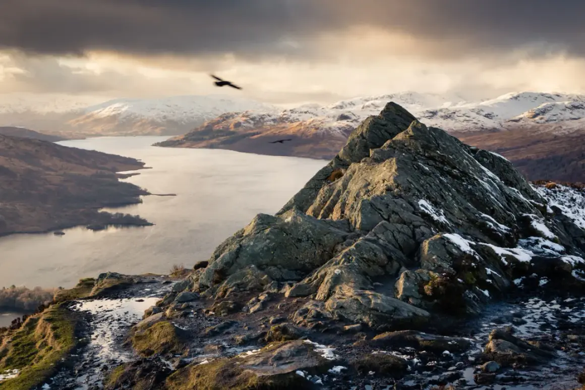 Vue spectaculaire d'une montagne écossaise enneigée, avec un lac et des oiseaux en vol.