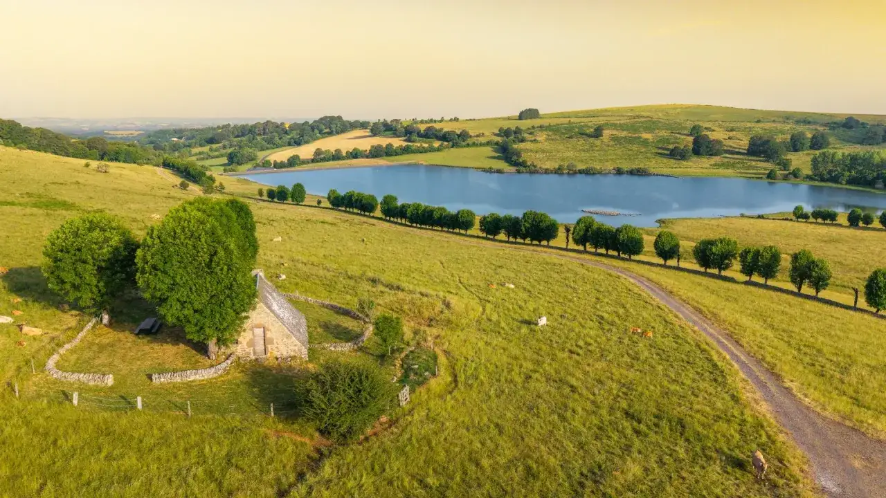 Vue aérienne du signal de Mailhebiau, avec un petit bâtiment en pierre, un lac et des collines verdoyantes.