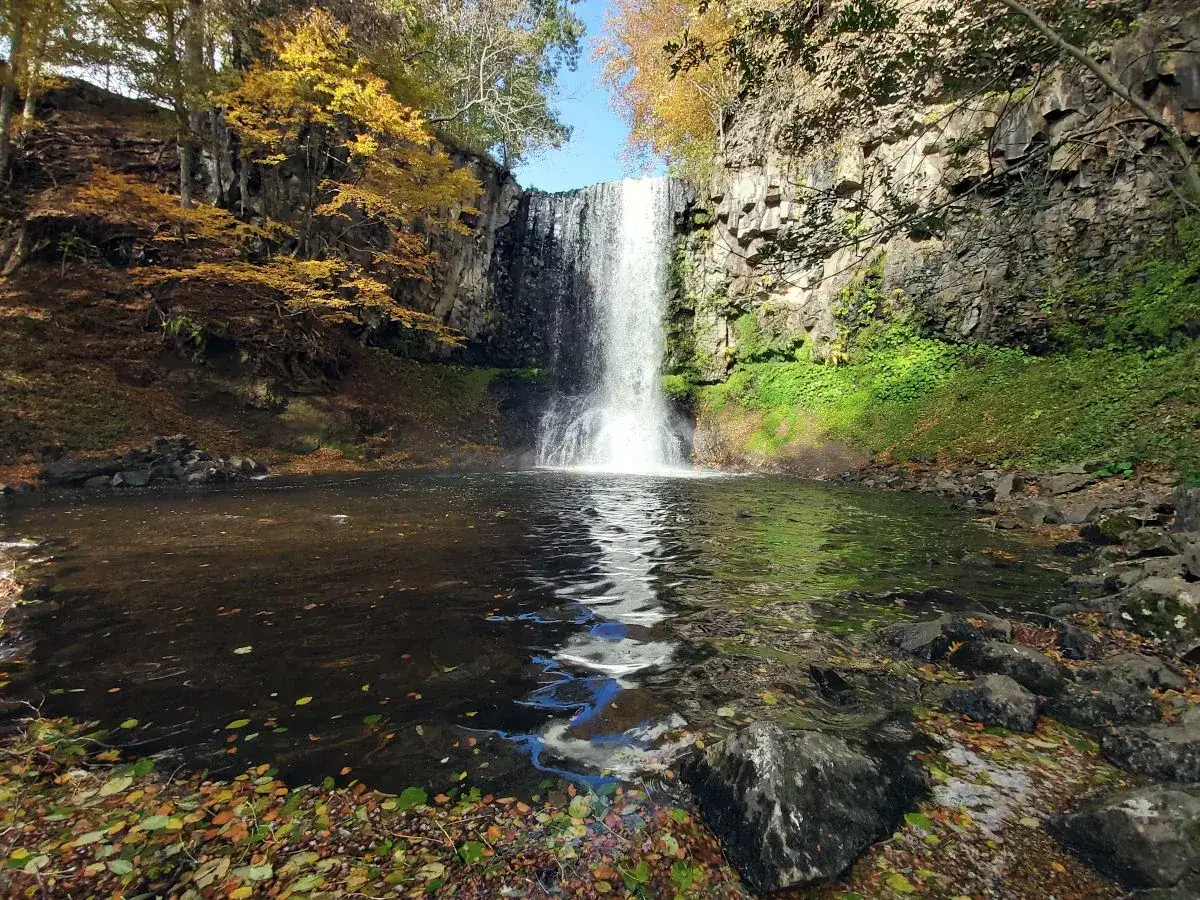 Cascade tombant dans un lac paisible, entouré de falaises rocheuses et d'arbres aux couleurs d'automne. Le lac d'Entraigues reflète le ciel bleu et la chute d'eau.