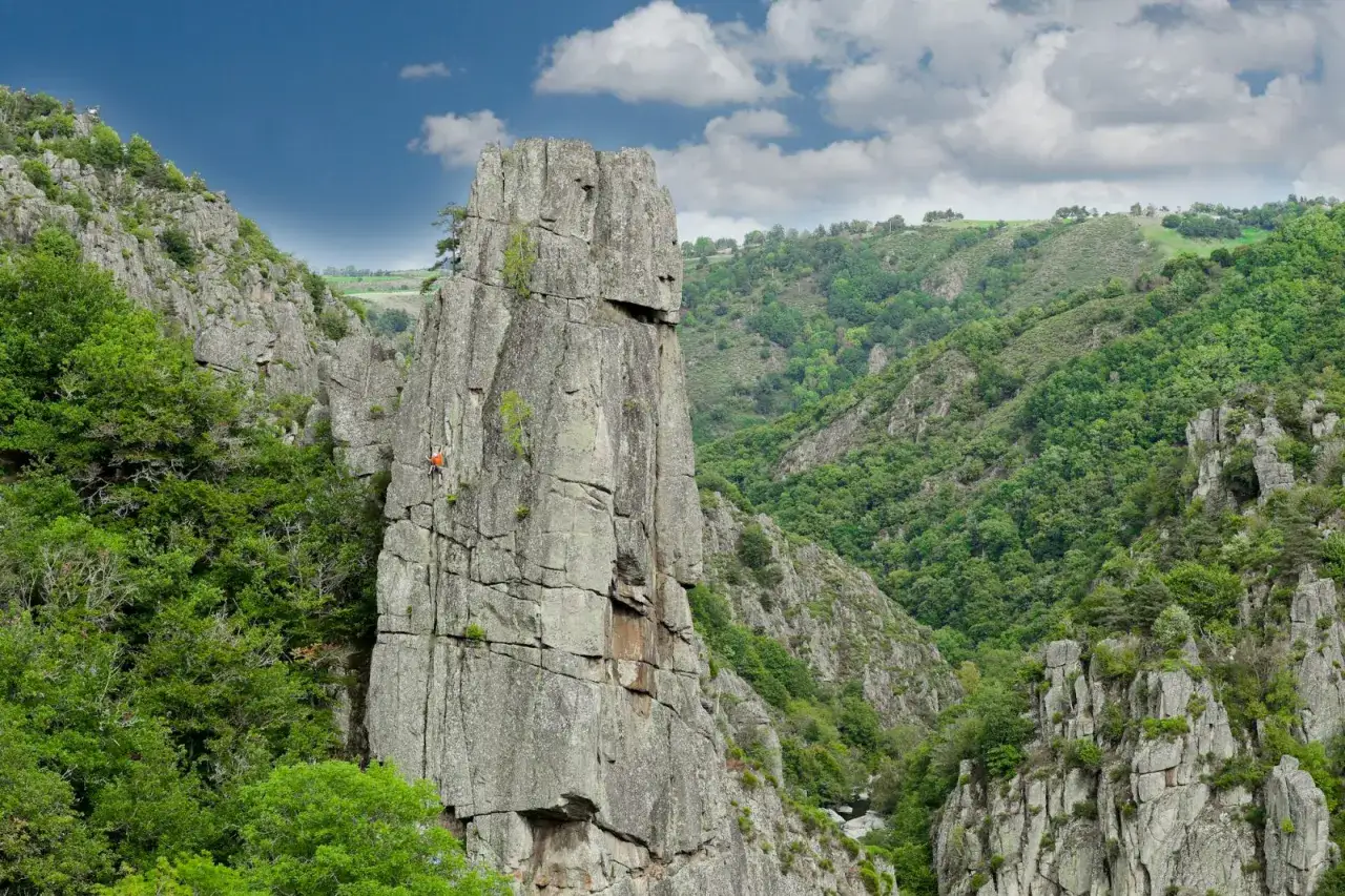 Escalade dans les gorges du Tarn, un grimpeur orange sur une paroi rocheuse imposante, entour&eacute;e de verdure luxuriante.
