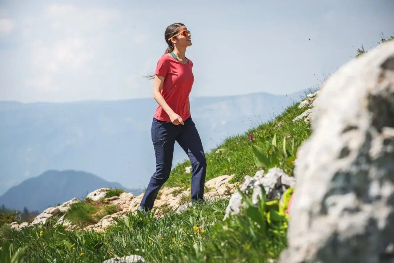 Une femme en tenue de sport, lunettes de soleil, profite d'une randonnée en montagne. Elle porte une chaussure sans chaussette, visiblement à l'aise.