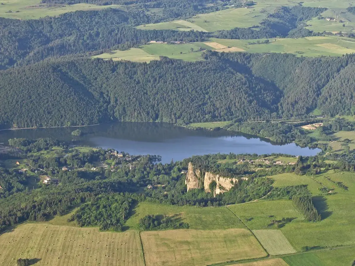 Vue aérienne d'un lac entouré de forêts et de champs. Un château médiéval en ruines domine un promontoire rocheux, idéal pour une randonnée à Murol.