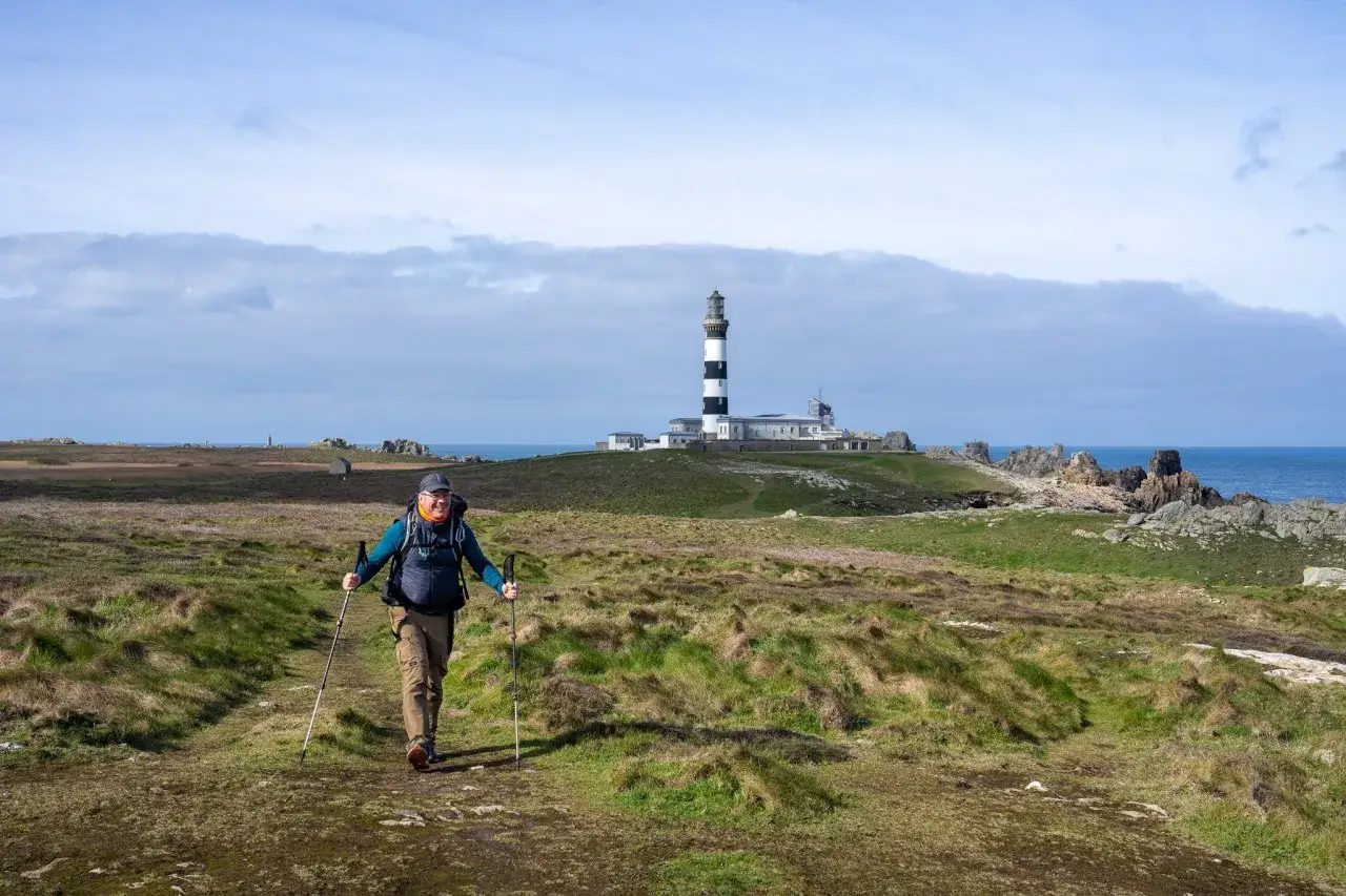 Un randonneur souriant marche sur un sentier côtier à Ouessant, le phare en arrière-plan.