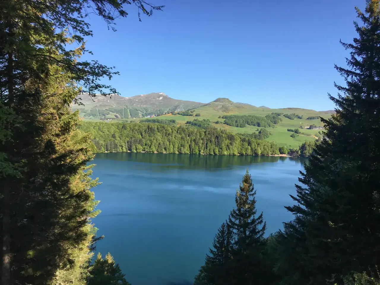 Vue panoramique sur un lac bleu profond, bordé d'une forêt verdoyante, avec en arrière-plan les montagnes du Puy de Sancy sous un ciel clair.