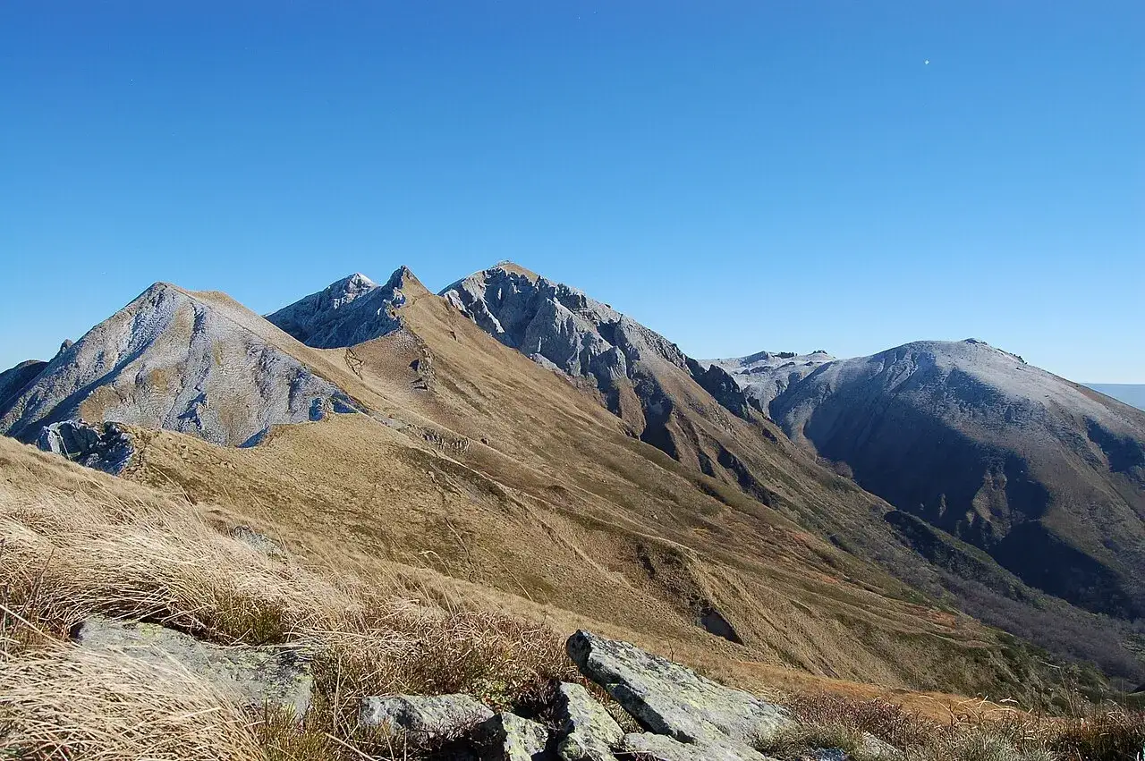 Vue panoramique des montagnes enneigées et des pentes herbeuses, évoquant un village autour du Mont-Dore sous un ciel bleu clair.