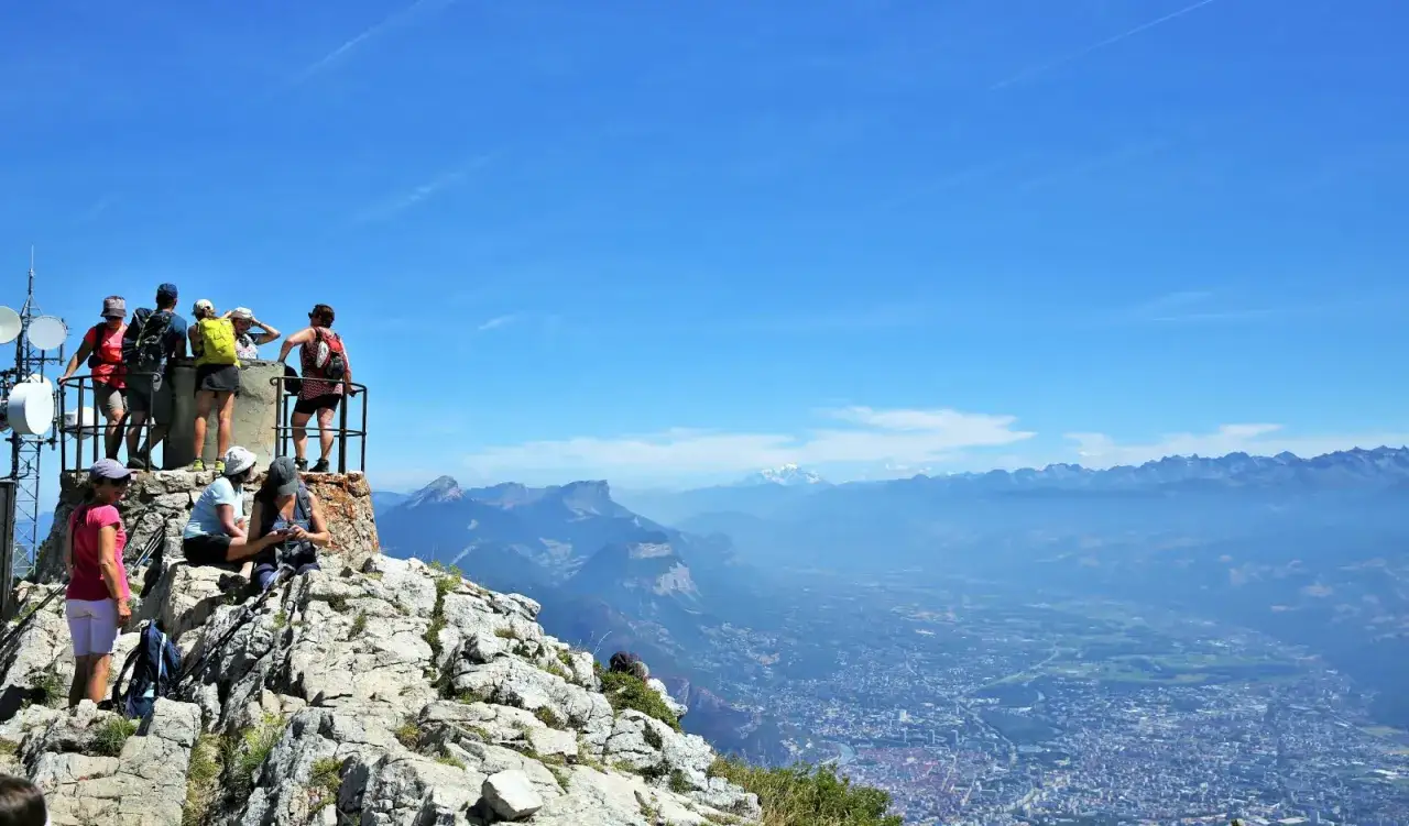 Des randonneurs admirent la vue depuis le sommet, un moucherotte perch&eacute; sur une antenne. La vall&eacute;e s'&eacute;tend &agrave; perte de vue sous un ciel bleu &eacute;clatant.