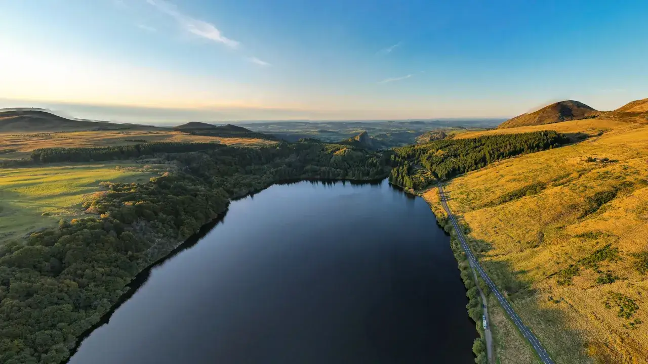 Vue aérienne du lac du Guéry au coucher du soleil, parfait pour une baignade paisible. Paysages verdoyants et collines dorées.