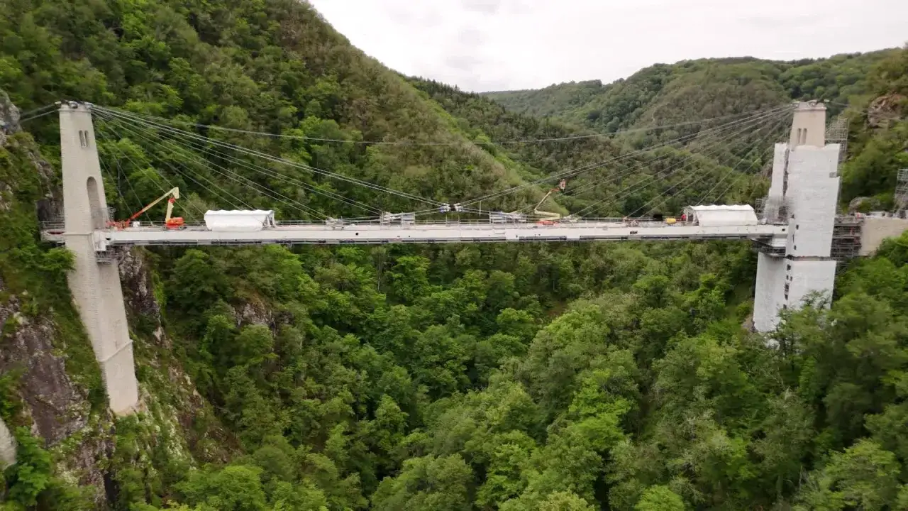 Le viaduc des Rochers Noirs, un pont suspendu impressionnant, traverse une vallée verdoyante. Des grues et des tentes de chantier indiquent des travaux en cours.