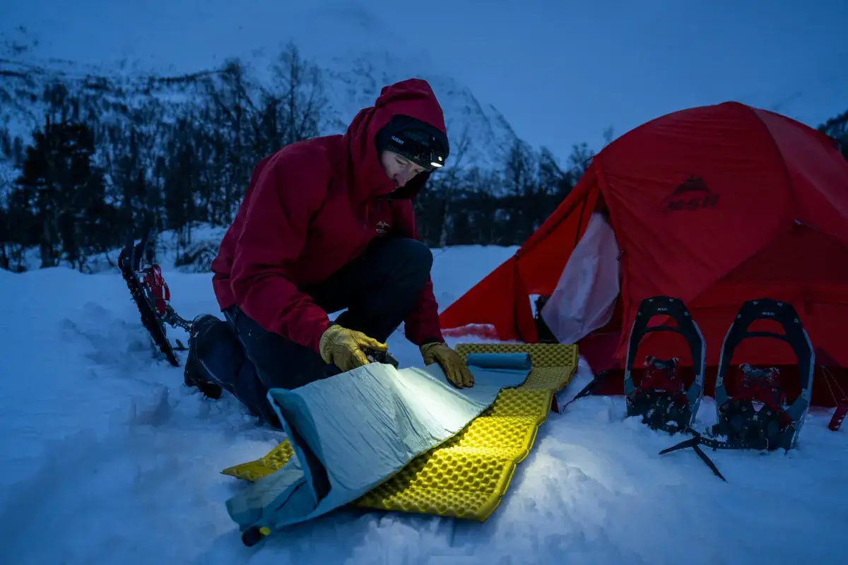 Pr&eacute;paration d'un campement hivernal. Une personne superpose deux matelas pour plus de confort. Raquettes et tente rouge &agrave; proximit&eacute;.