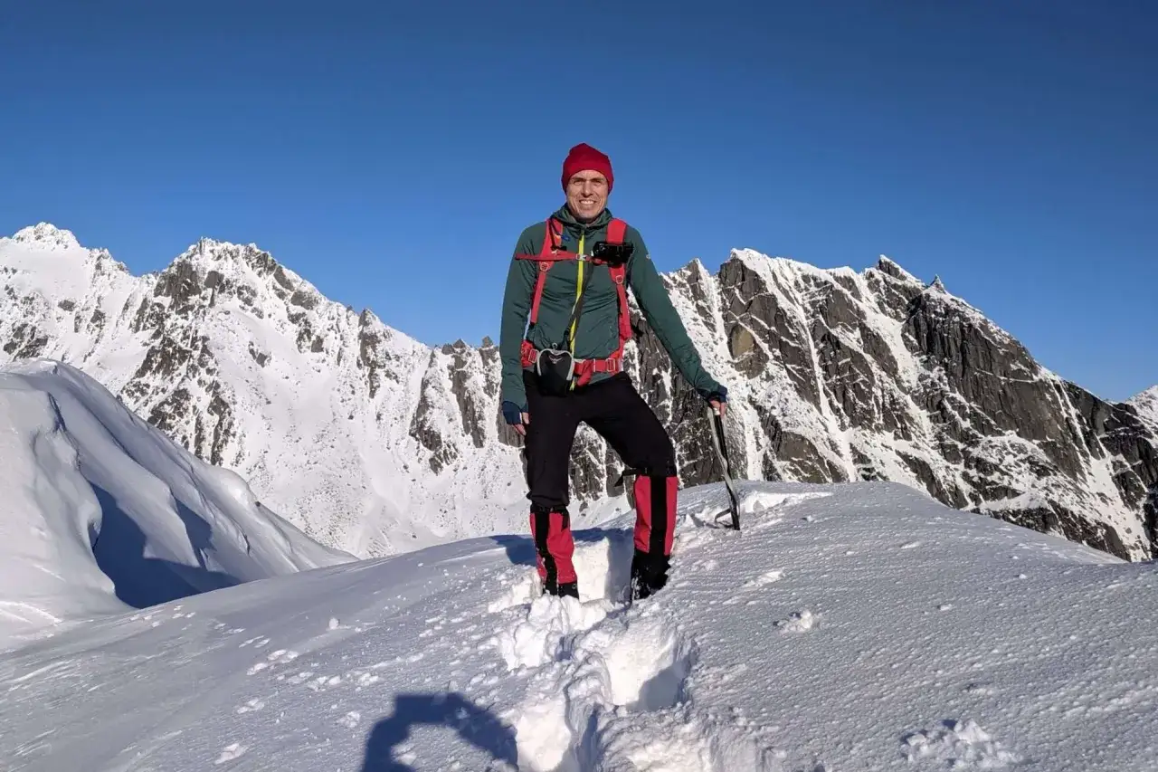 Un alpiniste souriant au sommet d'une montagne enneigée en Nouvelle-Zélande en septembre. Le ciel est d'un bleu éclatant.