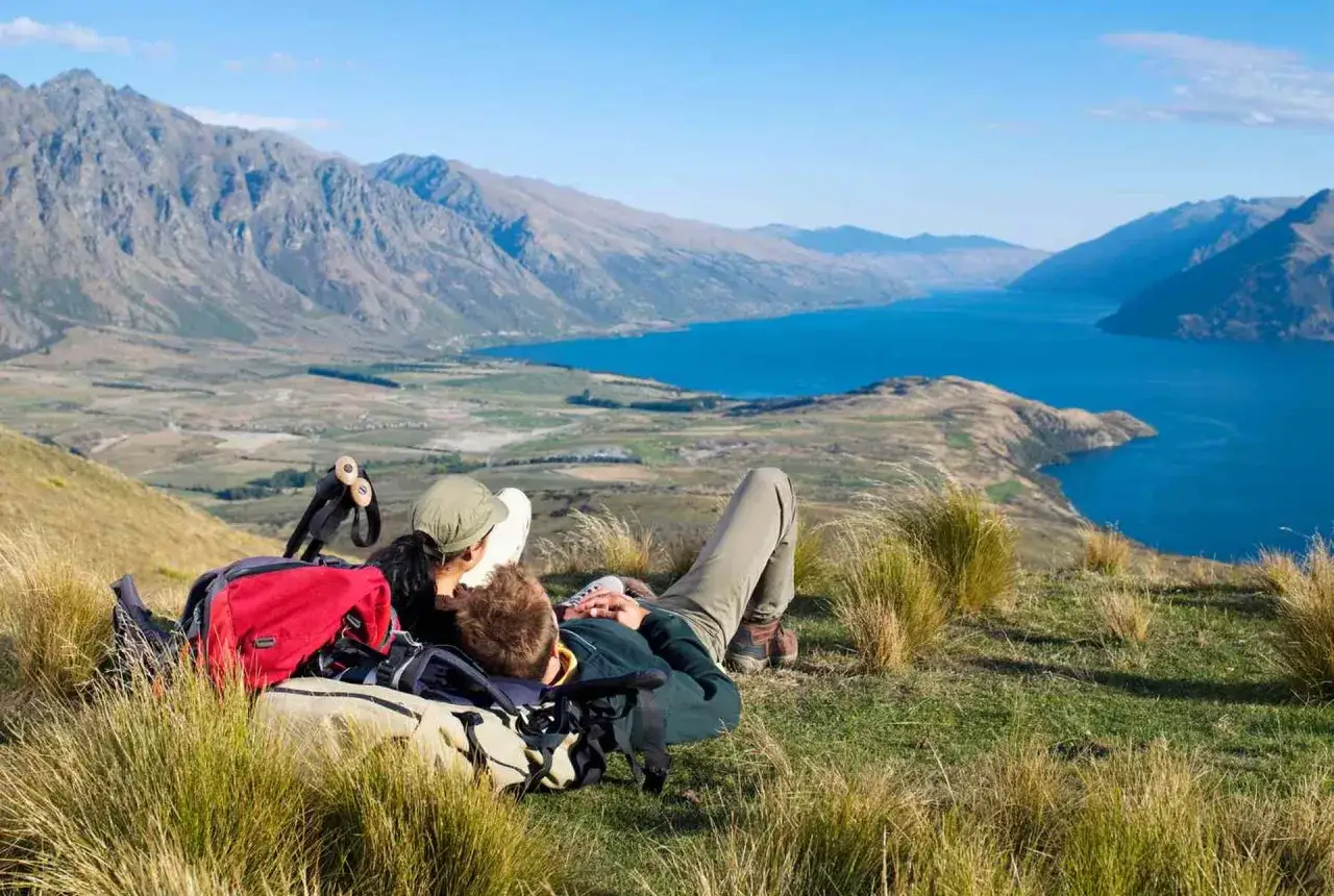 Deux randonneurs se reposent sur une colline en Nouvelle-Zélande, profitant d'une vue imprenable sur un lac et des montagnes. Un budget pour explorer ce paysage magnifique est une excellente idée.