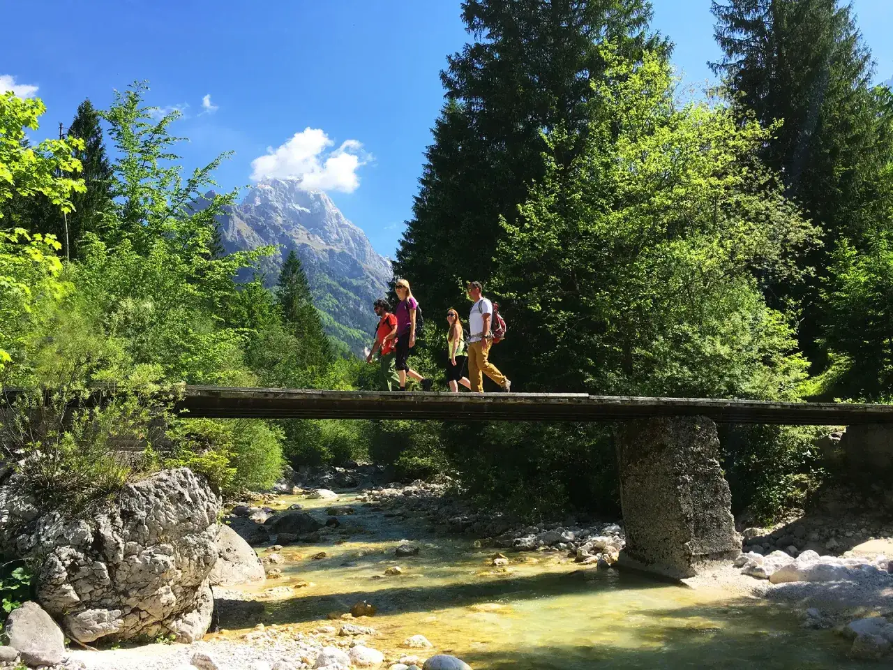 Randonneurs traversent un pont en bois sur une rivière turquoise, paysage de montagne en arrière-plan, sur l'Alpe Adria Trail.