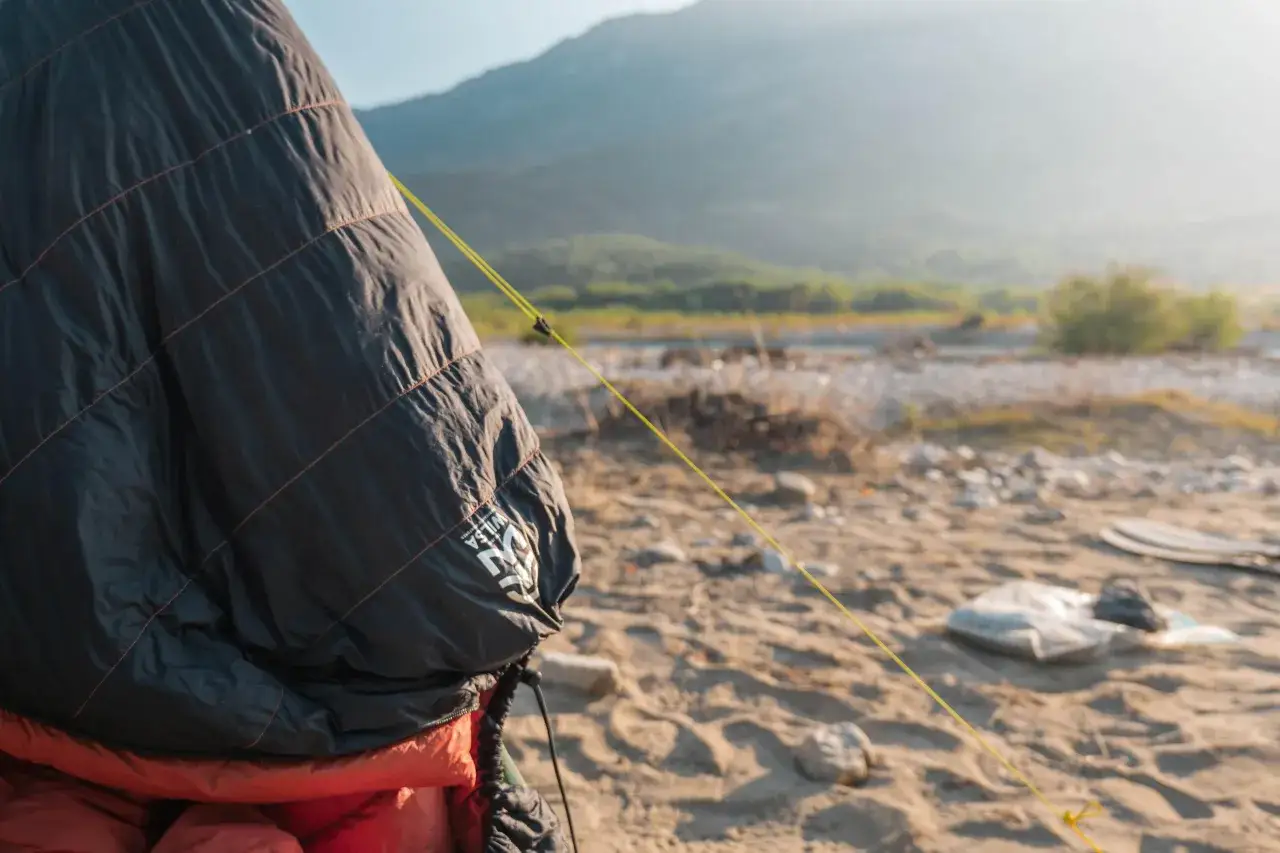 Sac de couchage noir et orange, pr&ecirc;t &agrave; &ecirc;tre rang&eacute;. Un fil jaune traverse le sable, rappelant comment ranger un sac de couchage pour le transport.
