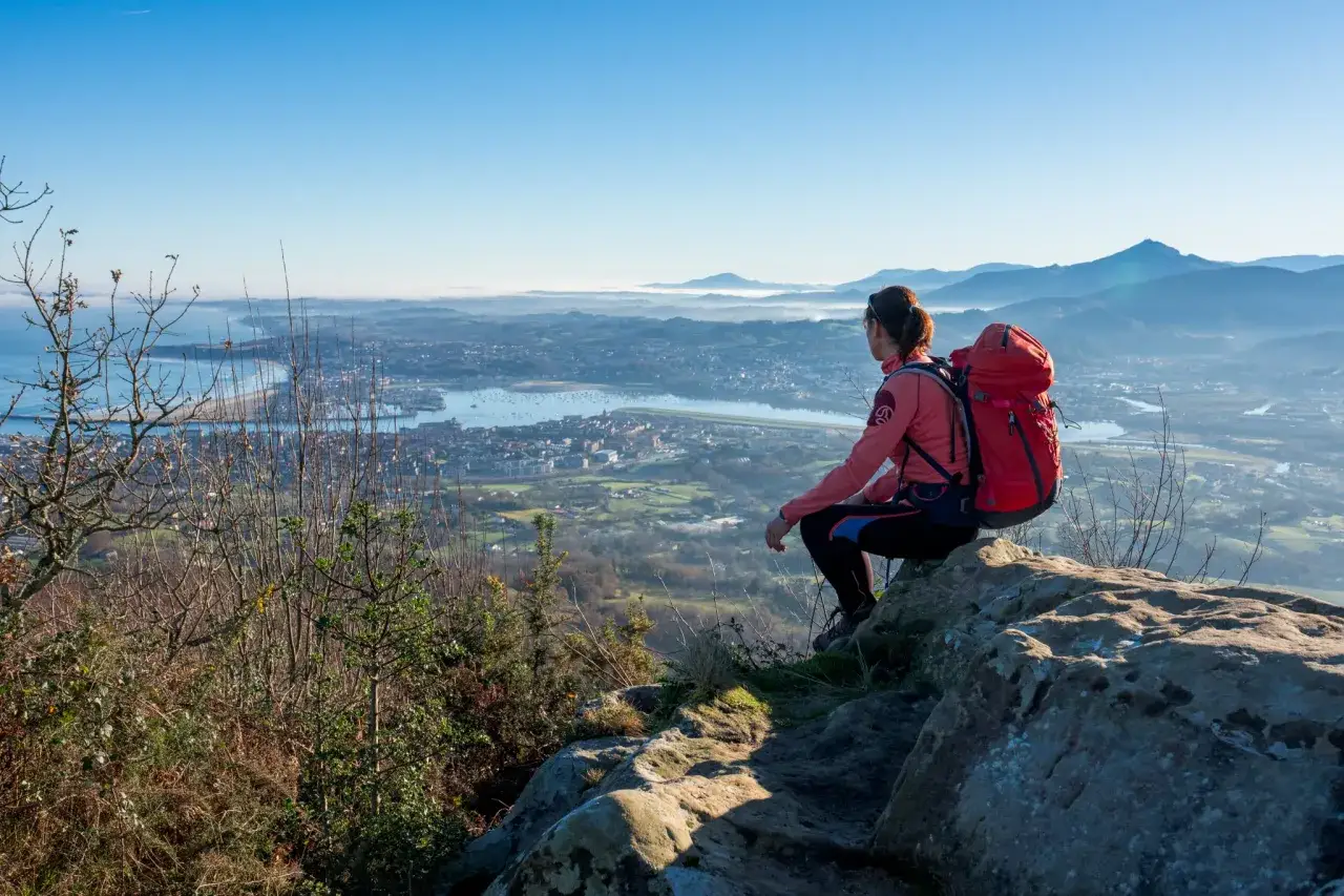 Vue panoramique de San Sebastián lors d'une randonnée. Une femme avec un sac à dos rouge admire le paysage depuis un sommet rocheux.