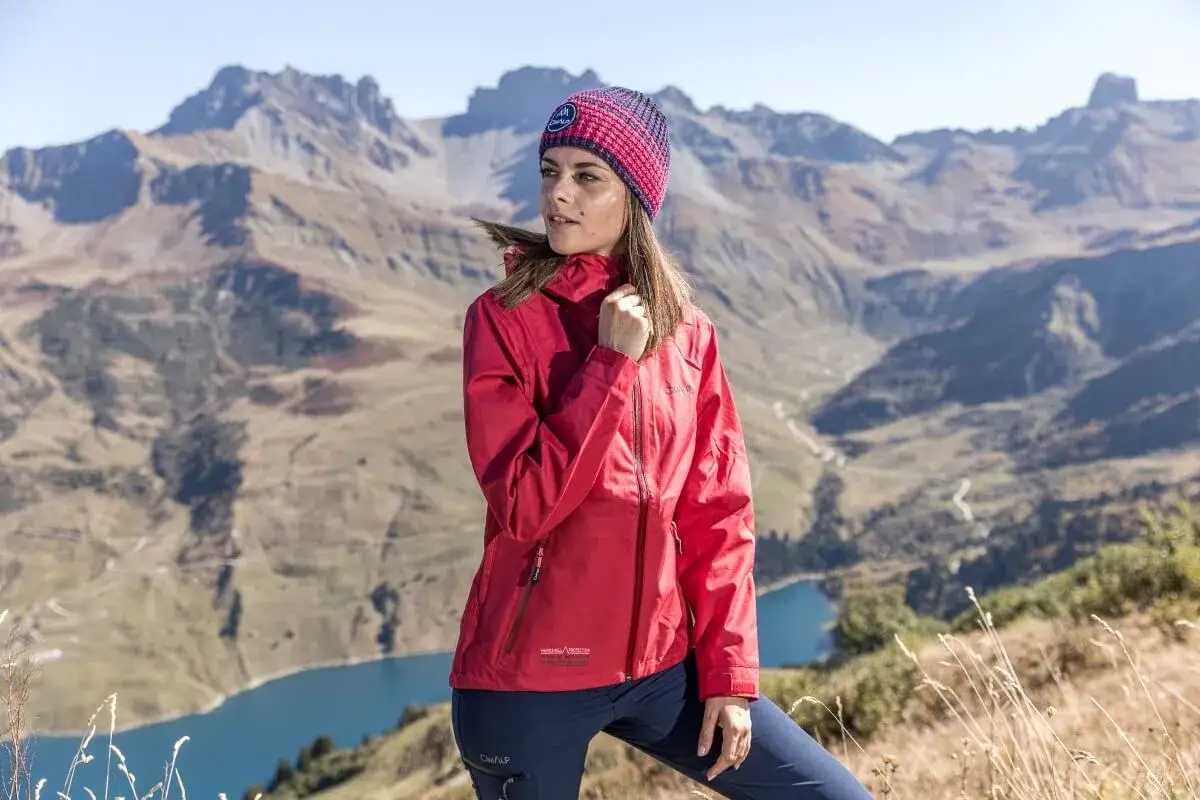 Femme en tenue de randonn&eacute;e rouge et bleue, sur fond de montagnes. Elle porte une veste qui illustre la diff&eacute;rence entre &eacute;tanche et imperm&eacute;able.