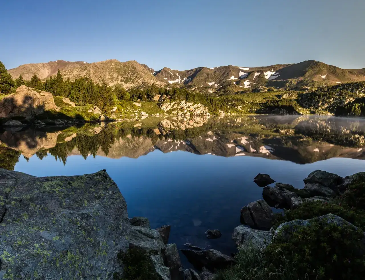 Matin calme au lac refl&eacute;tant les montagnes enneig&eacute;es du massif du Carlit. Rochers et pins bordent l'eau paisible.