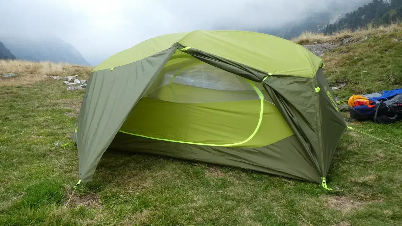 Tente verte et jaune, &eacute;quipement de camping Nemo, install&eacute;e sur une pelouse avec des montagnes brumeuses en arri&egrave;re-plan.