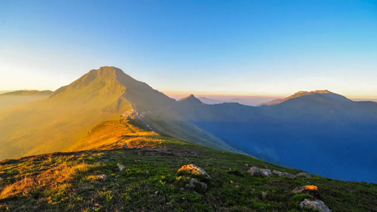 Le soleil dore les flancs du puy May, révélant une route sinueuse et des montagnes majestueuses sous un ciel clair.