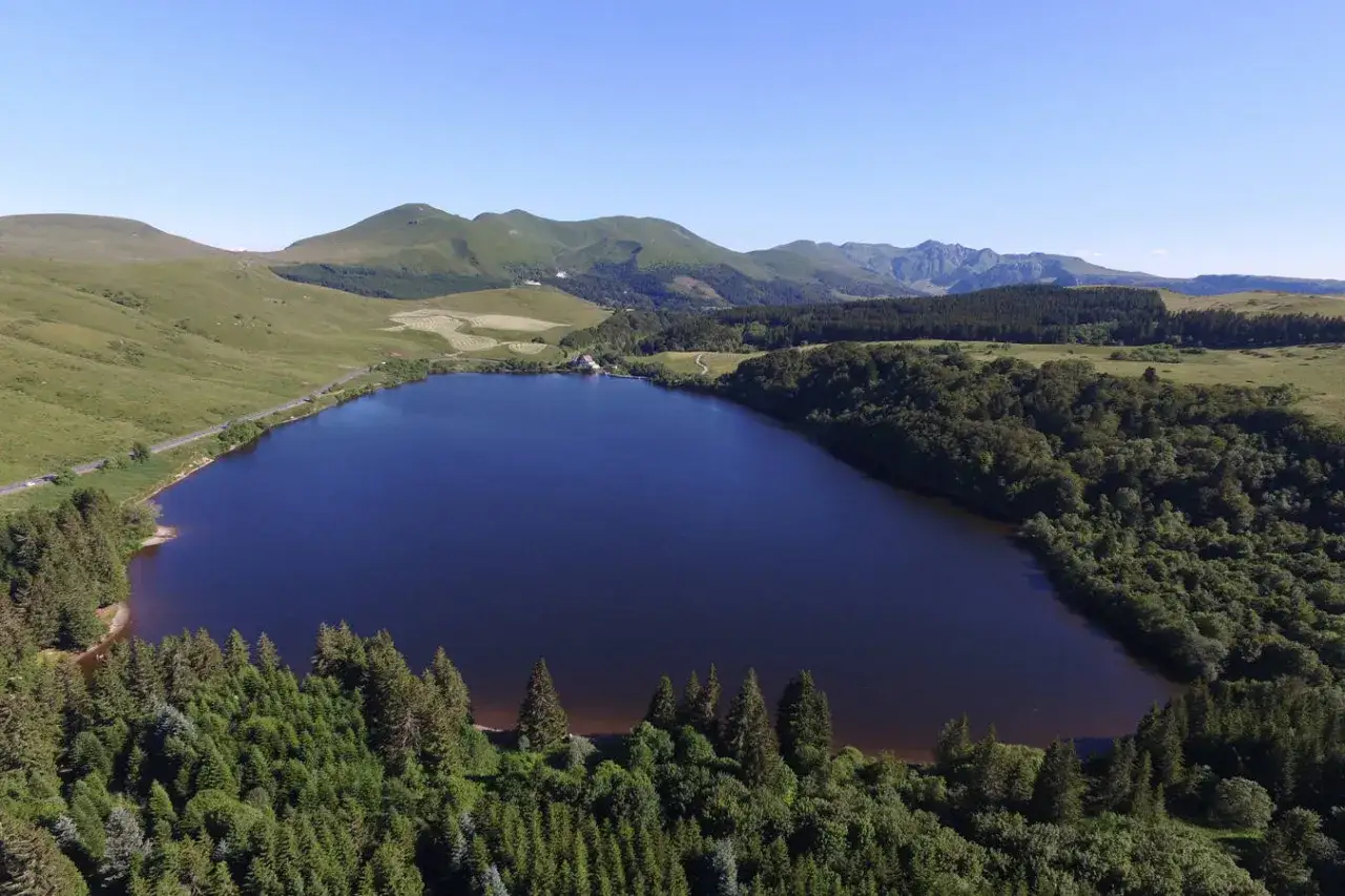 Vue aérienne du lac de Guéry, avec ses rives boisées et les montagnes en arrière-plan. Un itinéraire de randonnée serpente le long de la côte.