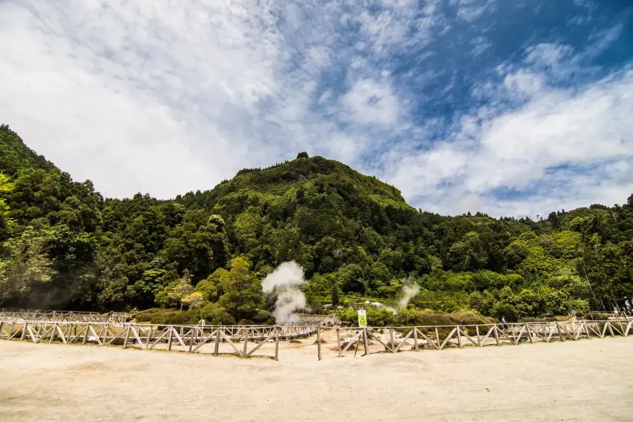 Vapeurs s'&eacute;chappant des fumerolles dans le paysage volcanique de Furnas, A&ccedil;ores, avec une montagne verdoyante en arri&egrave;re-plan.