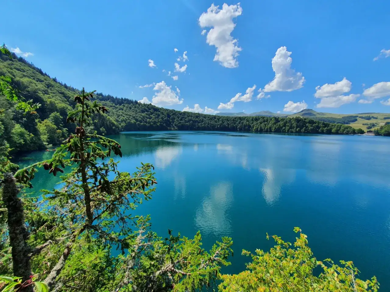 Lac d'un bleu profond, entouré de forêts verdoyantes et de collines douces sous un ciel d'été. Un paysage à voir en Auvergne.