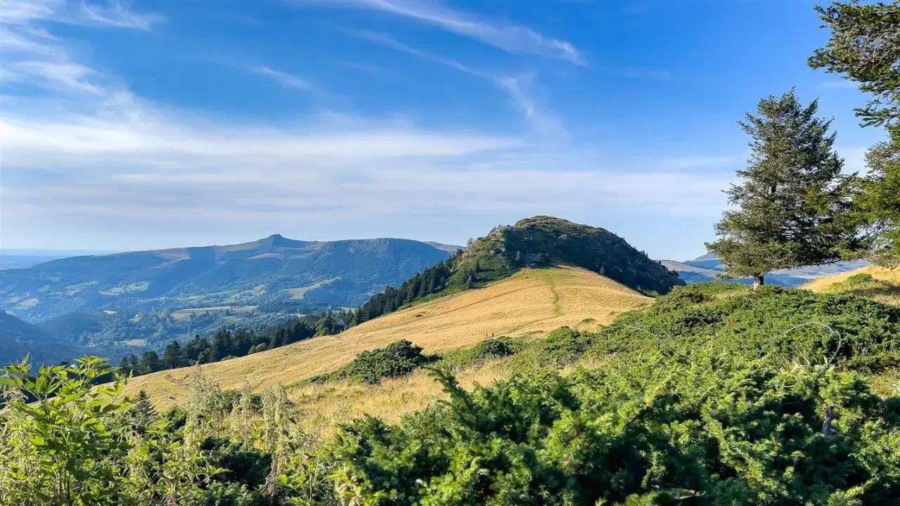 Vue panoramique d'une banne ordanche verdoyante sous un ciel bleu. Un sentier serpente sur la colline, menant vers des montagnes lointaines.