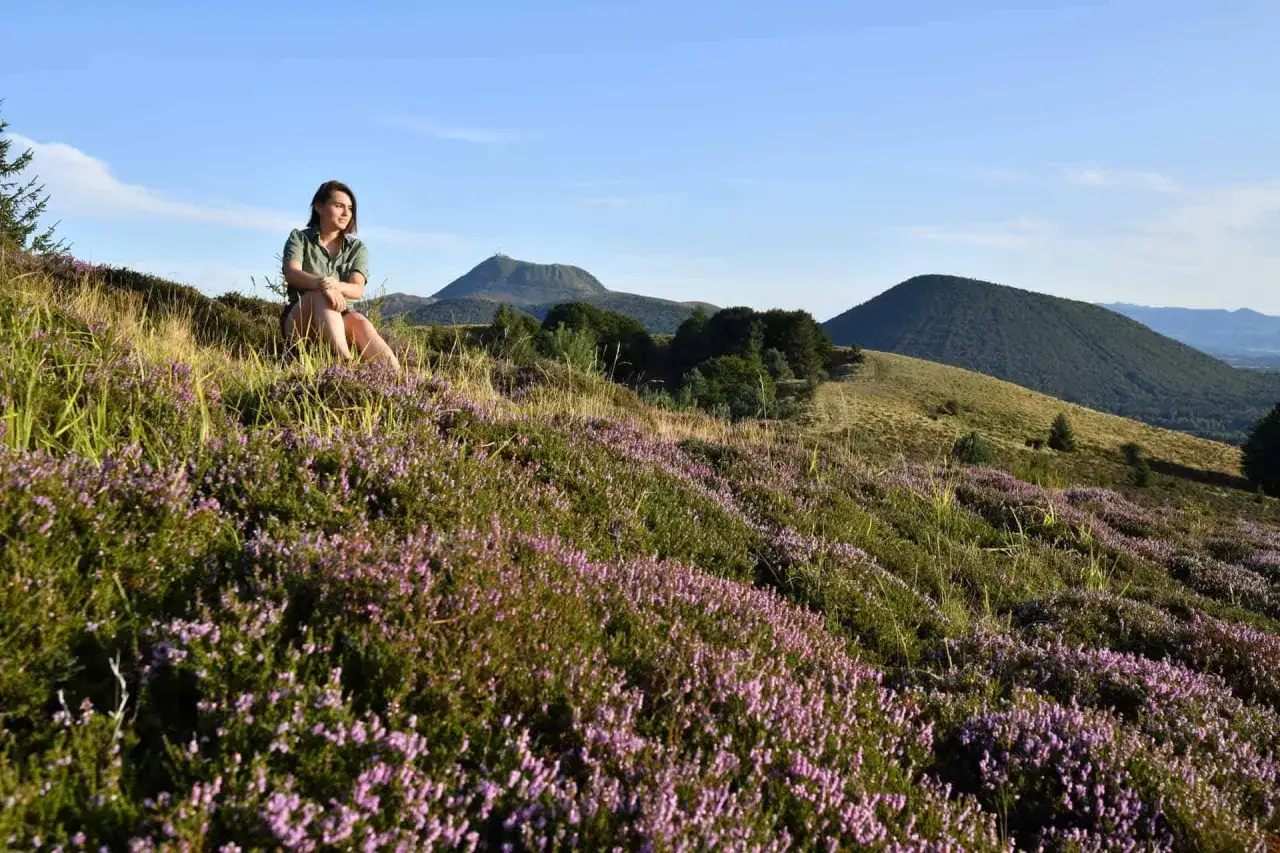 Jeune femme profitant d'une pause lors d'une rando dans la Chaine des Puys, entourée de bruyères en fleurs.