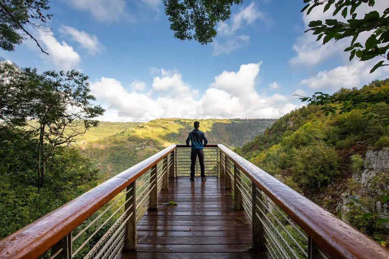Un randonneur admire le panorama des gorges de la Truyère depuis une passerelle en bois. Le ciel est bleu avec des nuages blancs.